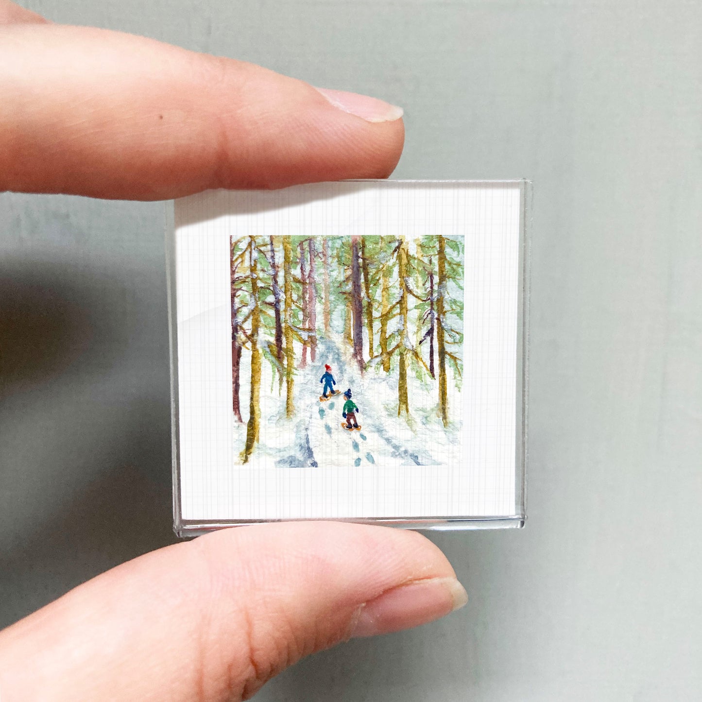 A person's hand is holding a small square picture depicting two people skiing through a snowy forest.