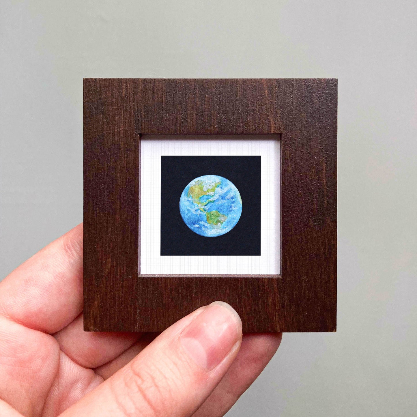 A hand is holding a small wooden frame containing a miniature representation of the Earth, with a black background and a white border.