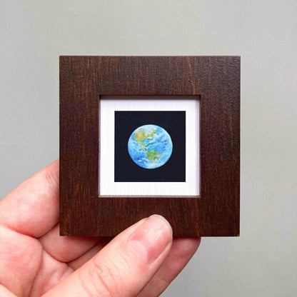 A hand is holding a small wooden frame containing a miniature representation of the Earth, with a black background and a white border.