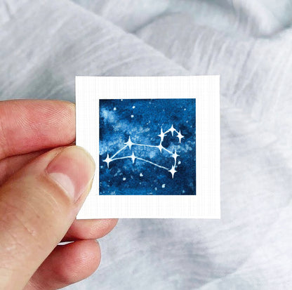 A hand is holding a small square-shaped paper with a depiction of a starry night sky on it.