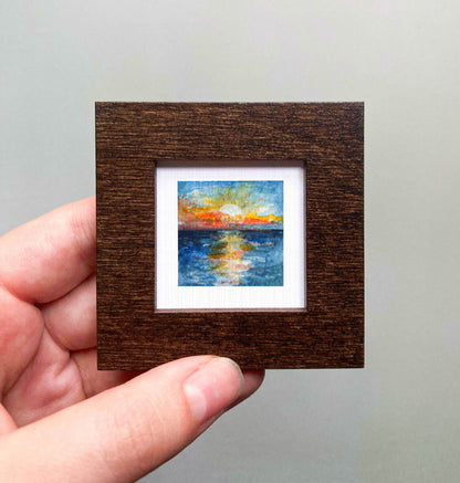 A hand is holding a small, square-shaped wooden frame containing a miniature painting of a sunset over a body of water.