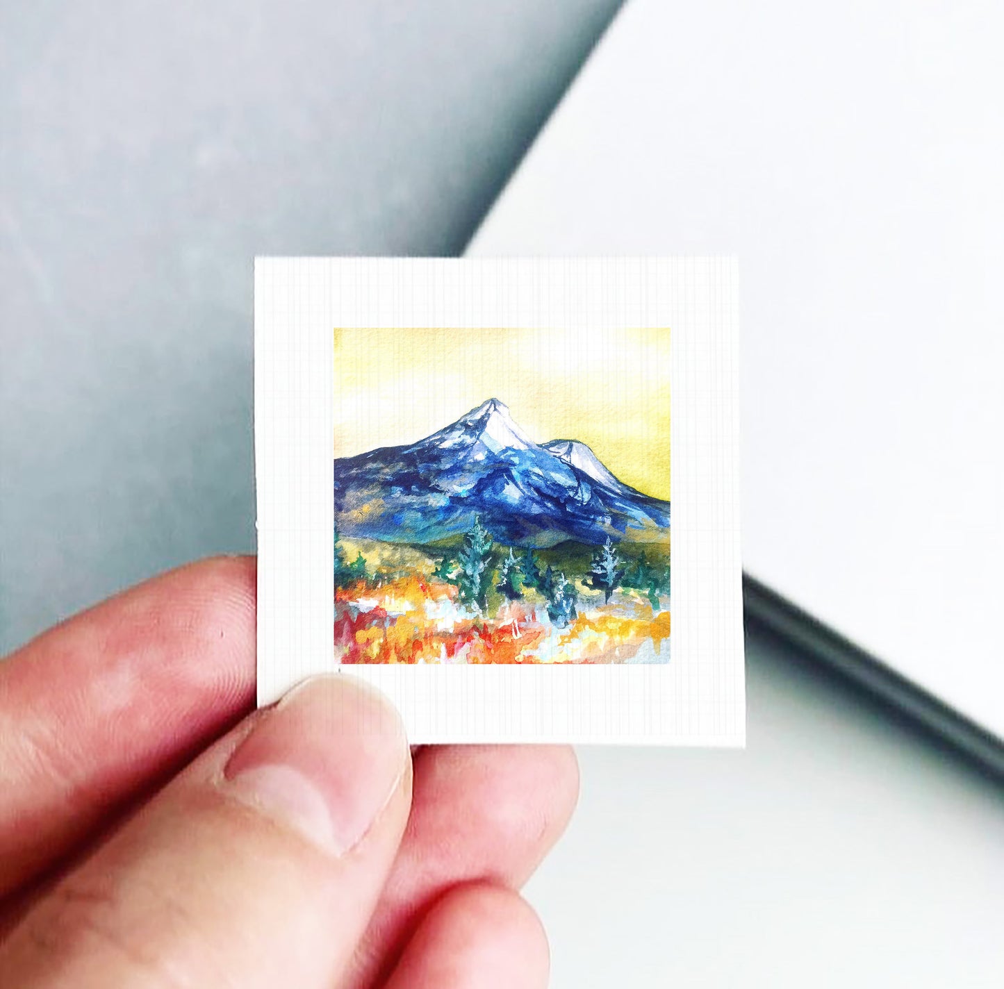 A hand is holding a small square artwork depicting a mountain landscape with a colorful, vibrant scene.