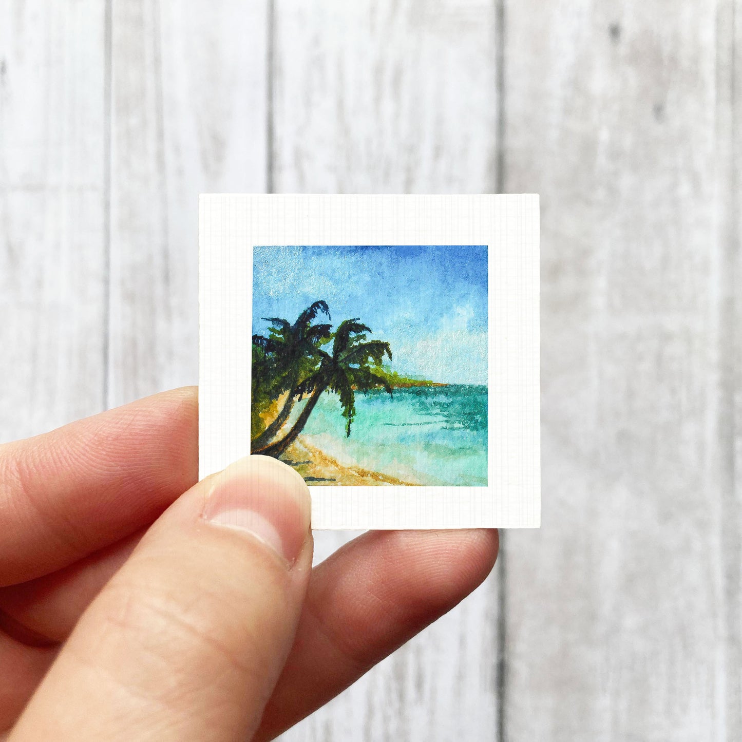 A hand is holding a small square painting of a tropical beach scene with palm trees and a blue sky.