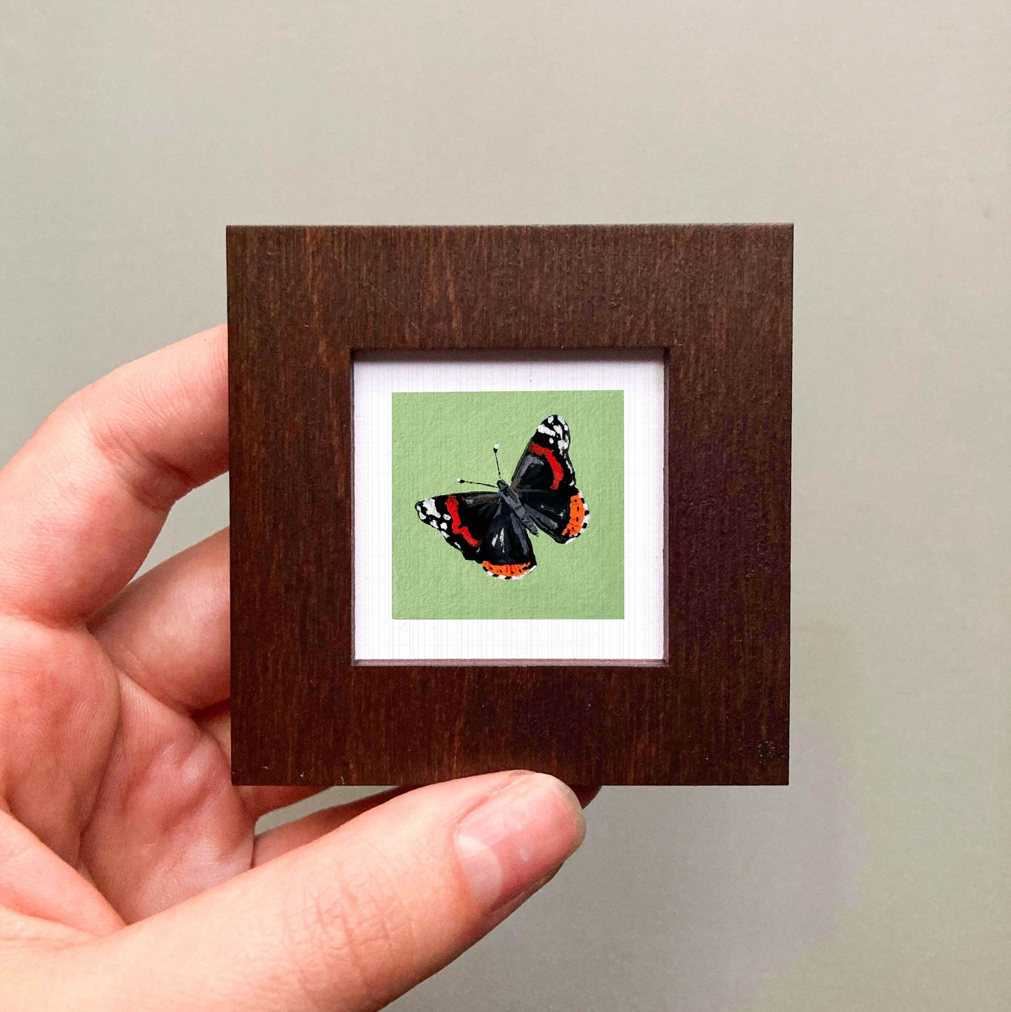 A hand is holding a small wooden frame containing a framed image of a butterfly.