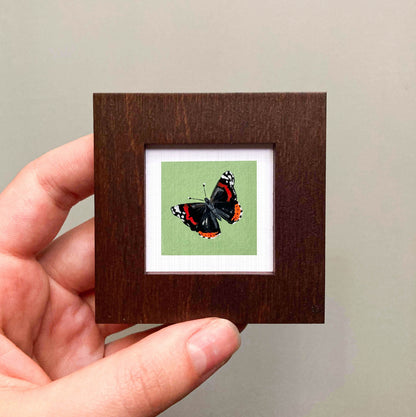 A hand is holding a small wooden frame containing a framed image of a butterfly.