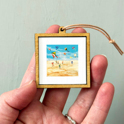 A person's hand is holding a small wooden frame containing a framed painting of people flying kites on a beach.
