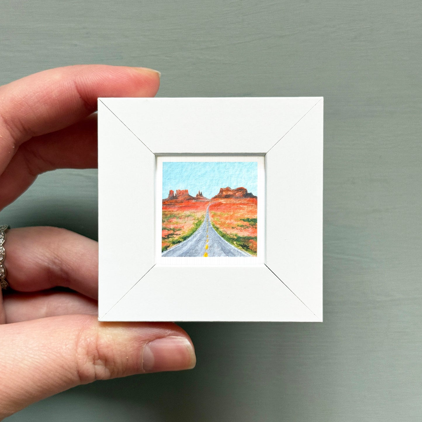 A hand is holding a small white square frame containing a miniature painting of a road leading to a desert landscape.
