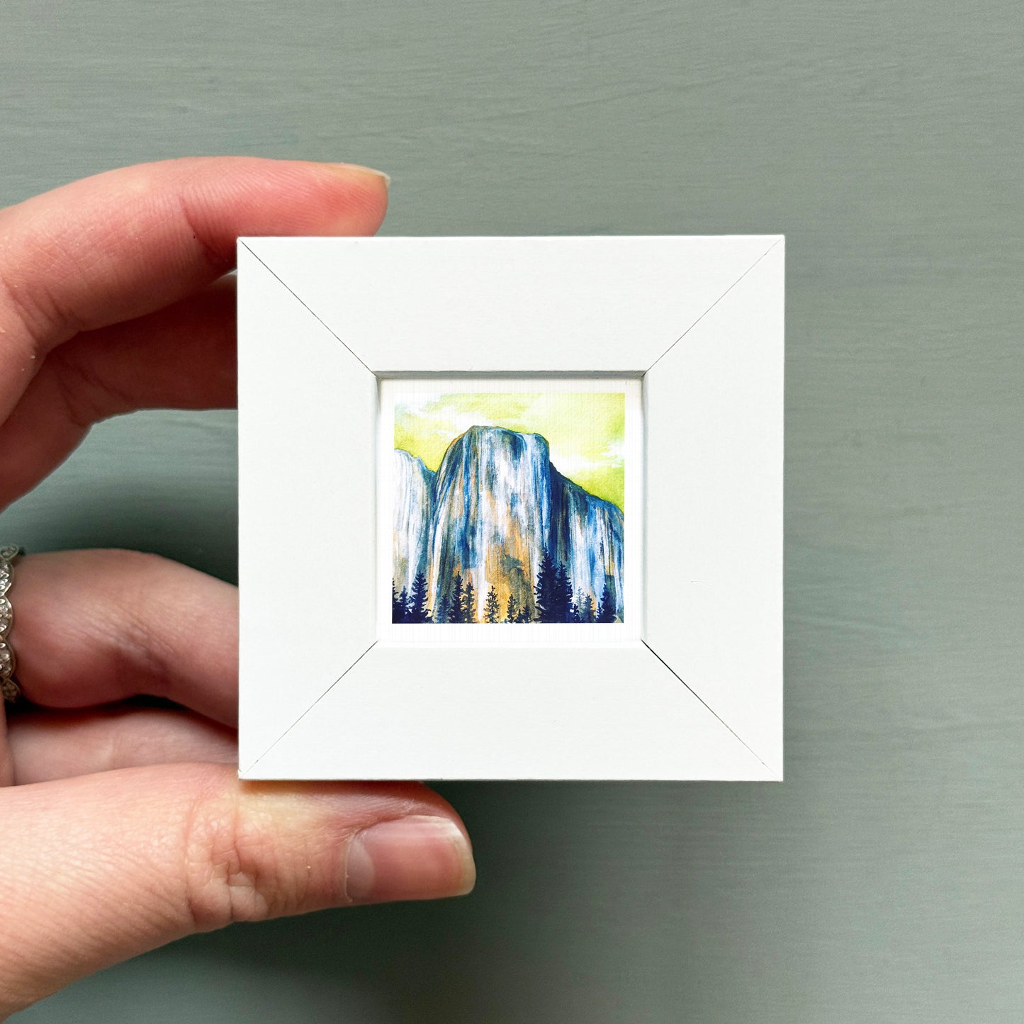 A hand is holding a small white square frame containing a painting of a mountain landscape.