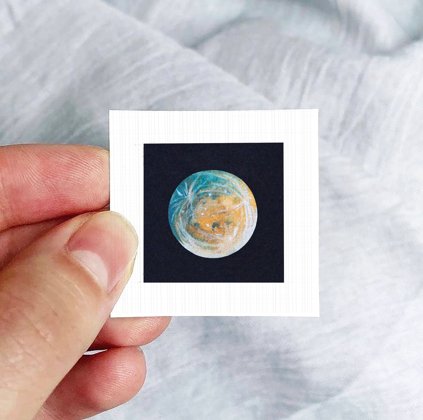 A hand is holding a small square photograph of a planet, which appears to be a representation of the Earth.