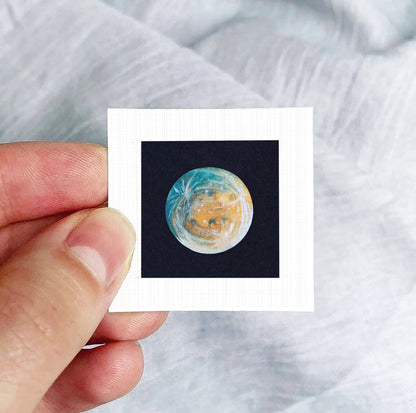 A hand is holding a small square photograph of a planet, which appears to be a representation of the Earth.