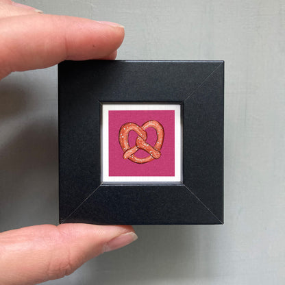 A hand is holding a small square frame containing a framed image of a pretzel.