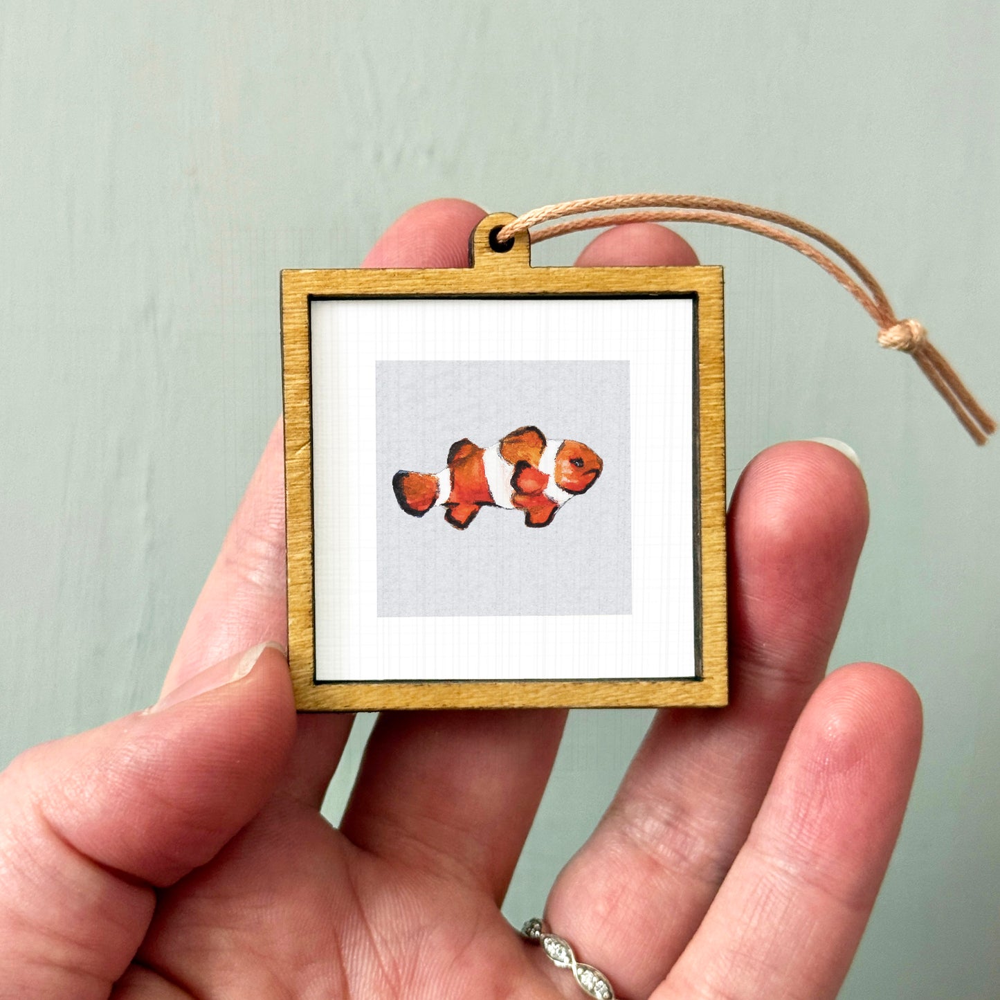A person's hand holding a small wooden frame containing a picture of a clownfish.