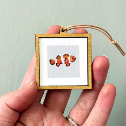 A person's hand holding a small wooden frame containing a picture of a clownfish.