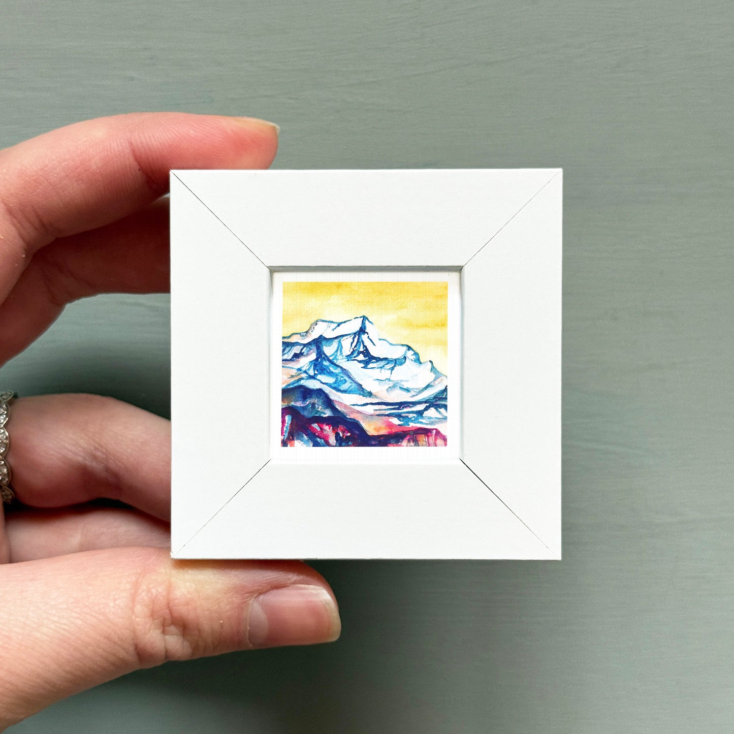 A hand is holding a small white square frame containing a watercolor painting of a mountain landscape.