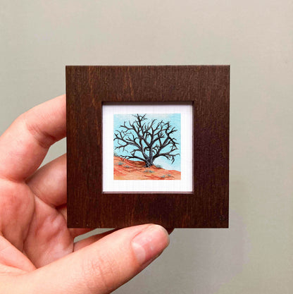 A hand is holding a small, square-shaped wooden frame containing a painting of a tree with bare branches against a blue sky.