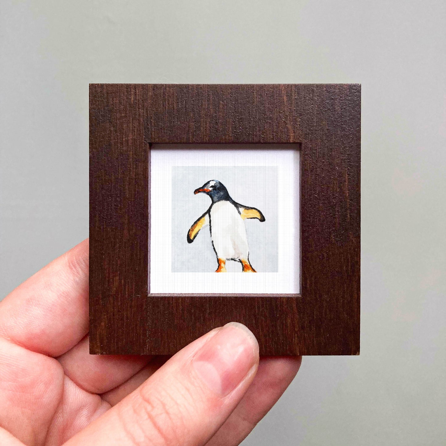 A hand is holding a small wooden frame containing a picture of a penguin.