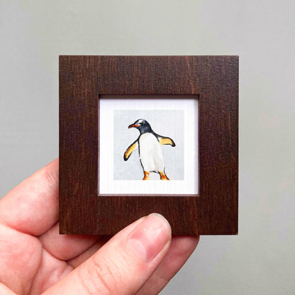 A hand is holding a small wooden frame containing a picture of a penguin.