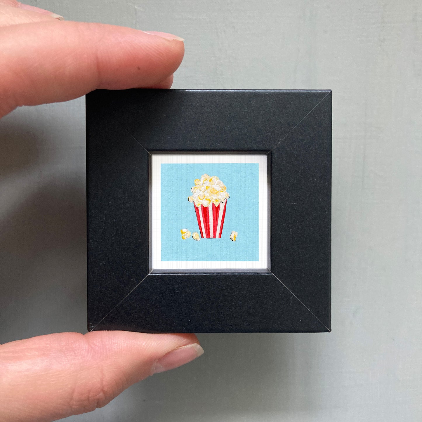 A hand is holding a small black picture frame containing a vibrant illustration of a red and white striped popcorn container with popcorn spilling out.