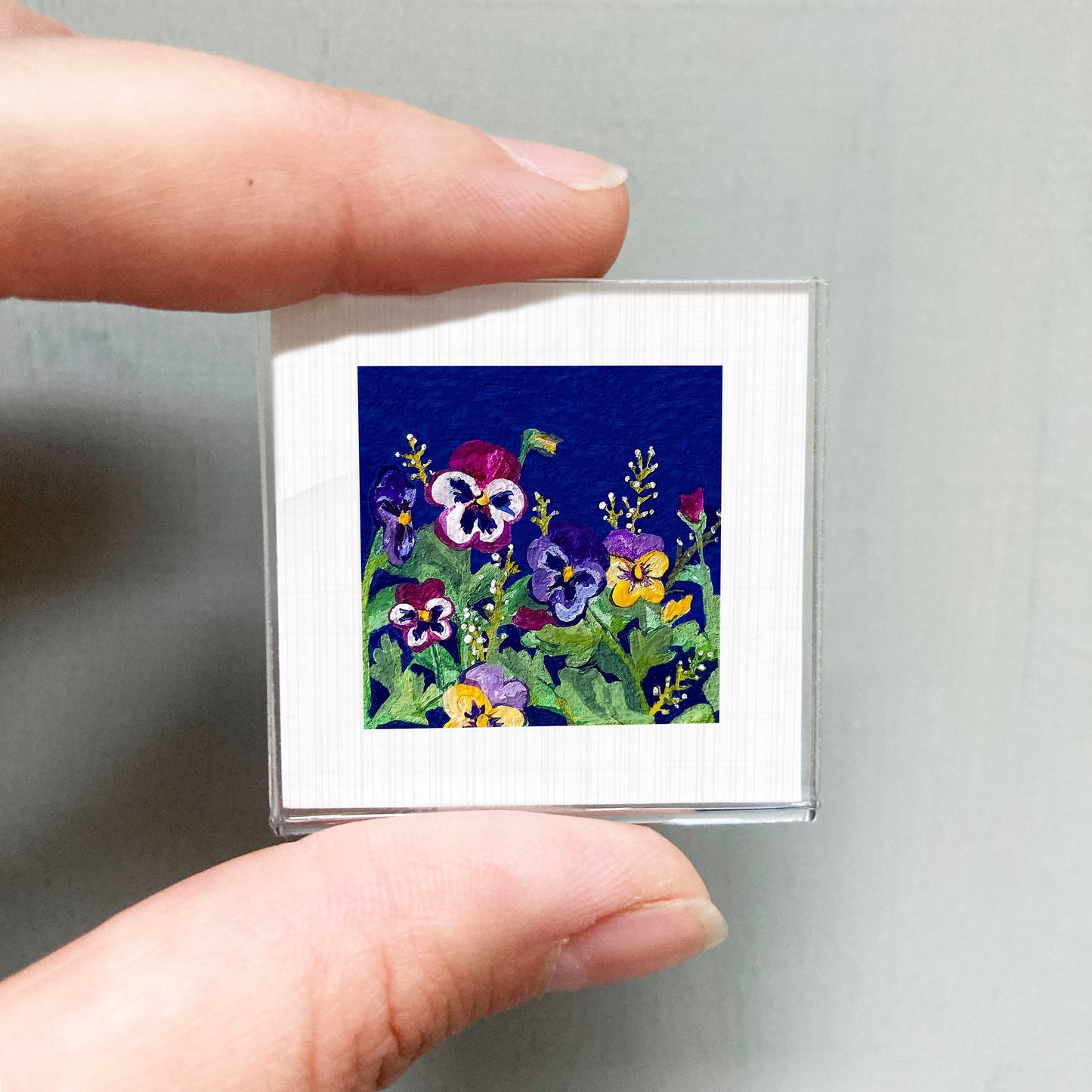A person's hand is holding a small square picture of a colorful floral scene.