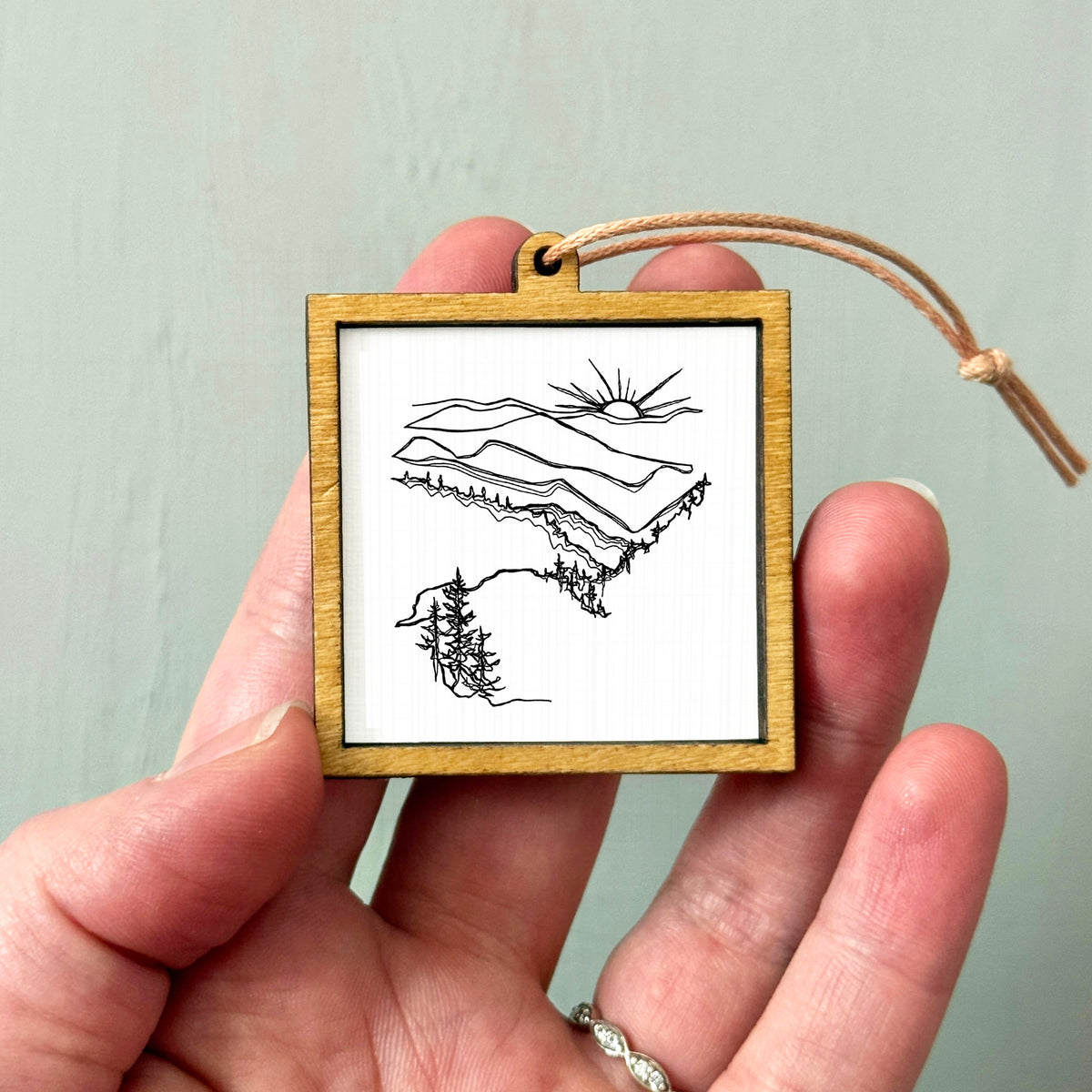 A hand holding a small wooden frame containing a black and white line drawing of a mountain landscape.