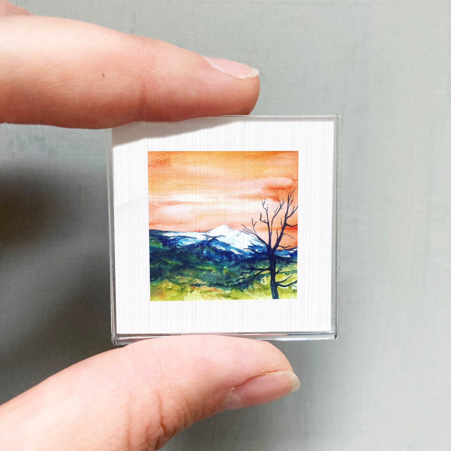 A person's hand holding a small square picture of a landscape with mountains and a tree.