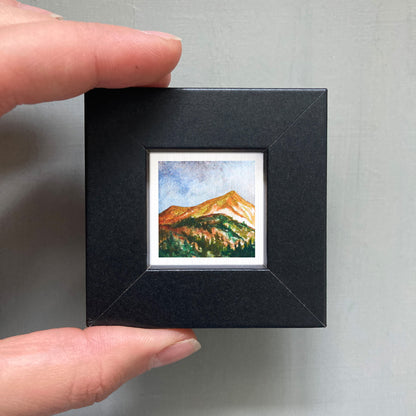 A person's hand holding a small, square-shaped black frame containing a watercolor painting of a mountain landscape.