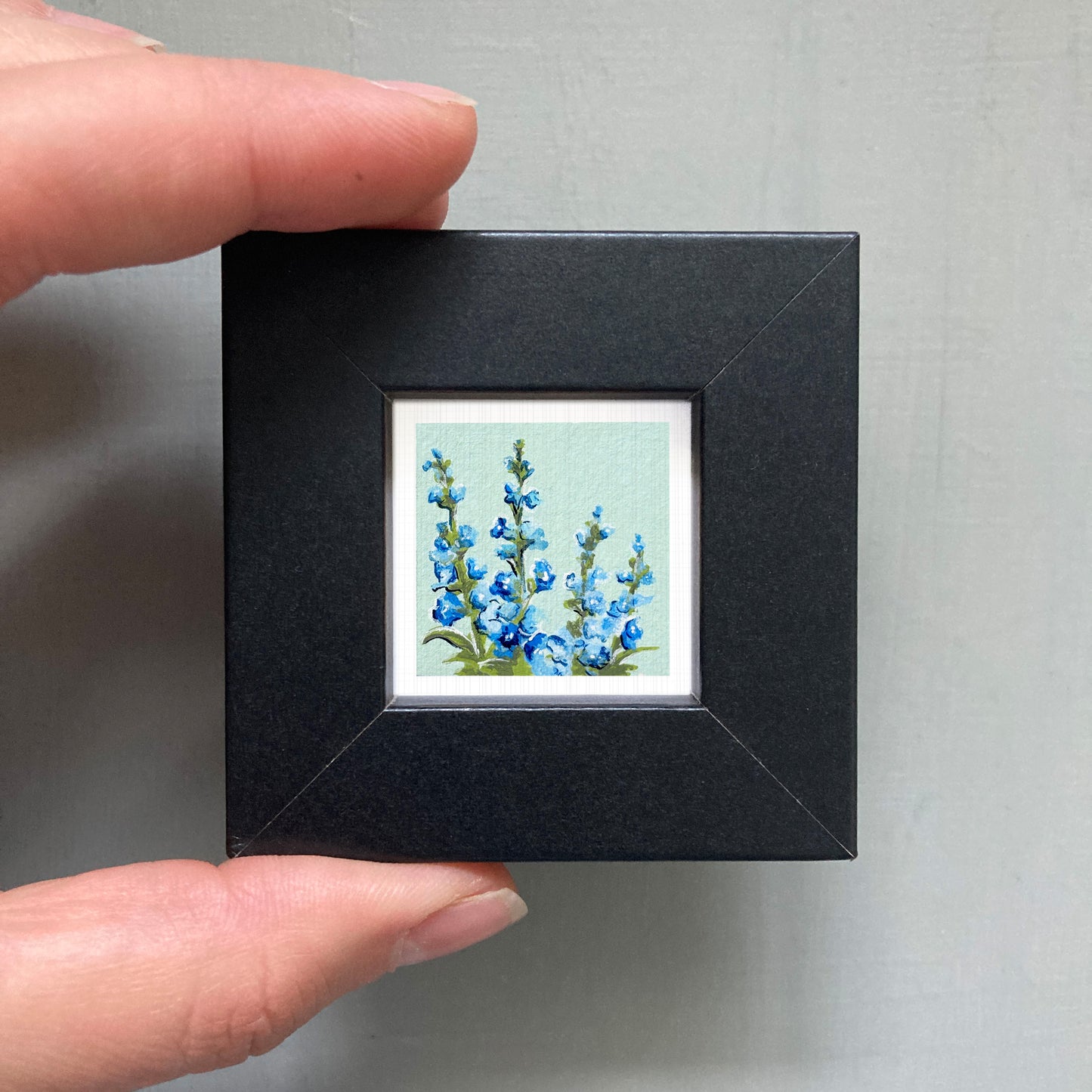 A person's hand holding a small black picture frame containing a framed painting of blue flowers.