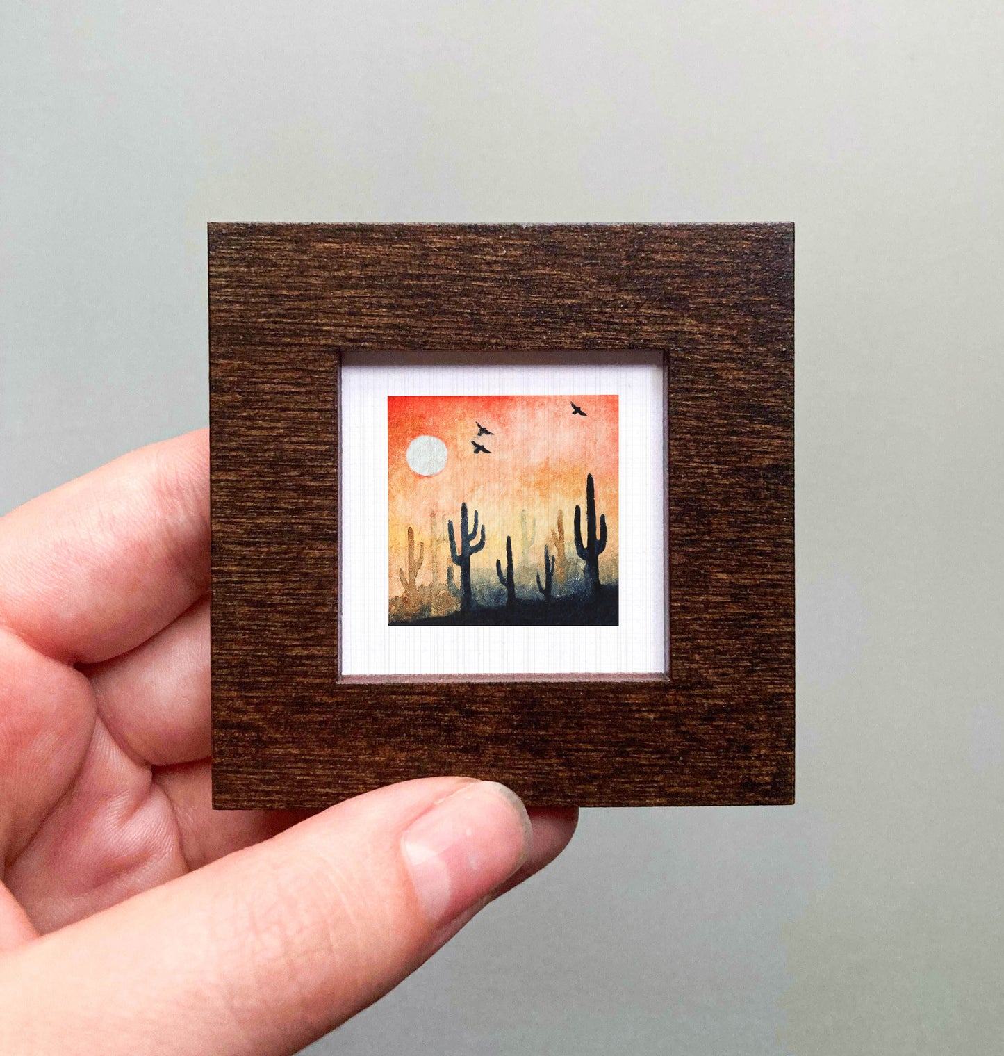 A hand is holding a small framed painting of a desert landscape with cacti and a full moon in the sky.