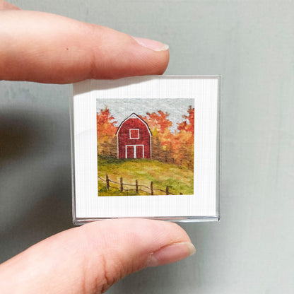 A person's hand holding a small square picture of a red barn surrounded by autumn foliage.