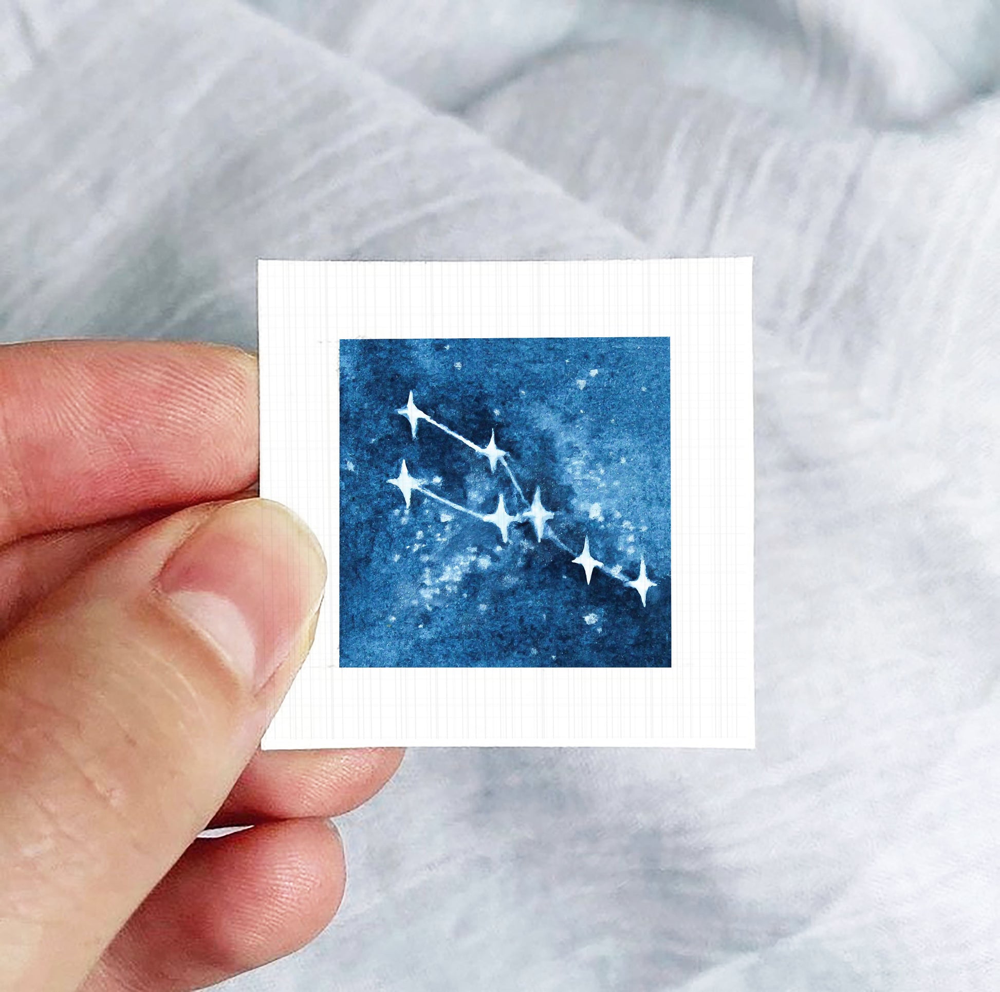 A hand is holding a small square-shaped paper with a blue starry night sky image on it.