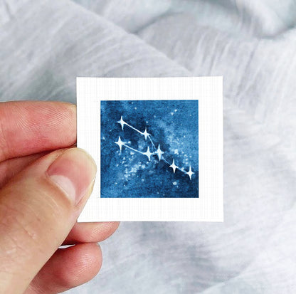 A hand is holding a small square-shaped paper with a blue starry night sky image on it.