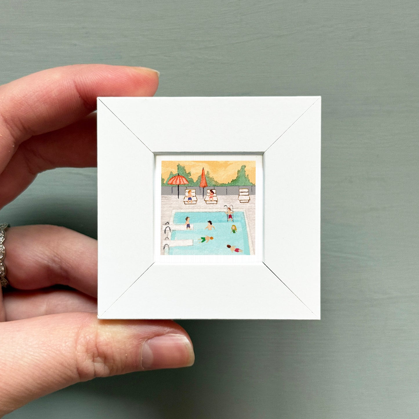 A hand is holding a small white picture frame containing an illustration of people swimming in a pool.