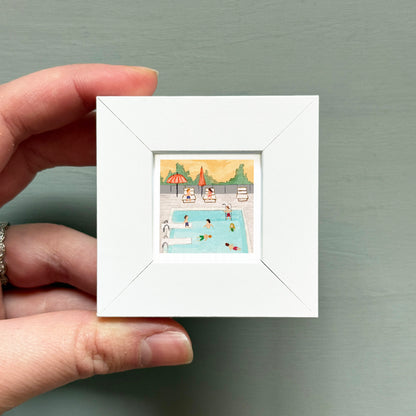 A hand is holding a small white picture frame containing an illustration of people swimming in a pool.