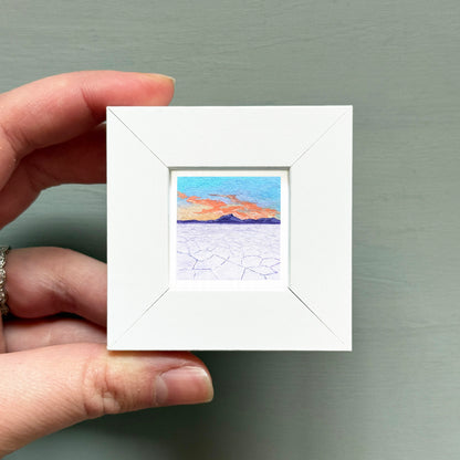 A hand is holding a small white picture frame containing a painting of a desert landscape with a sunset in the background.