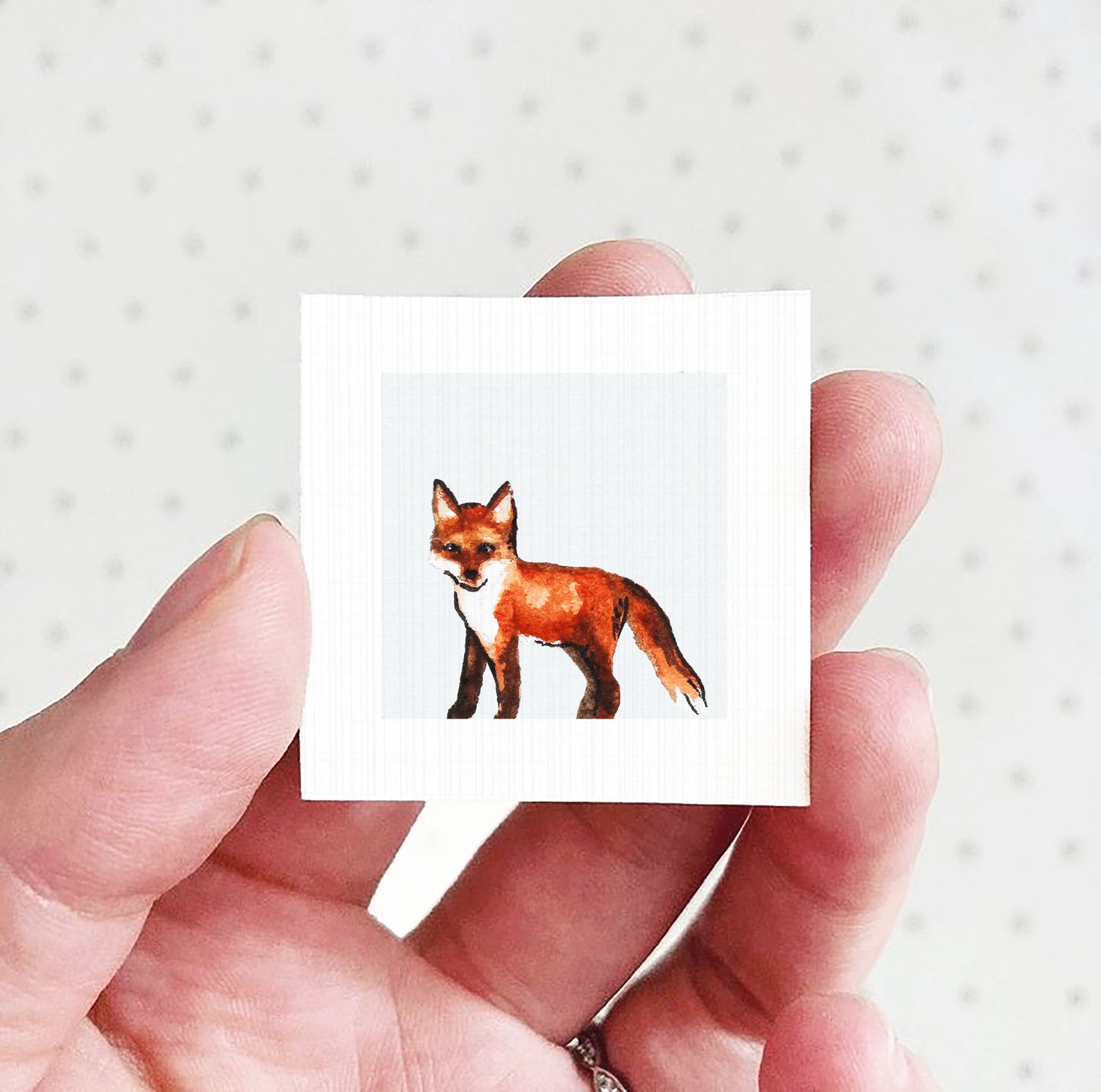 A hand holding a small square picture of a fox against a white background.