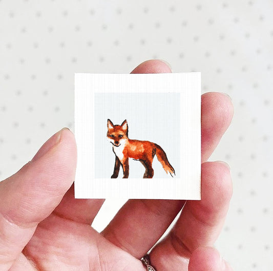 A hand holding a small square picture of a fox against a white background.