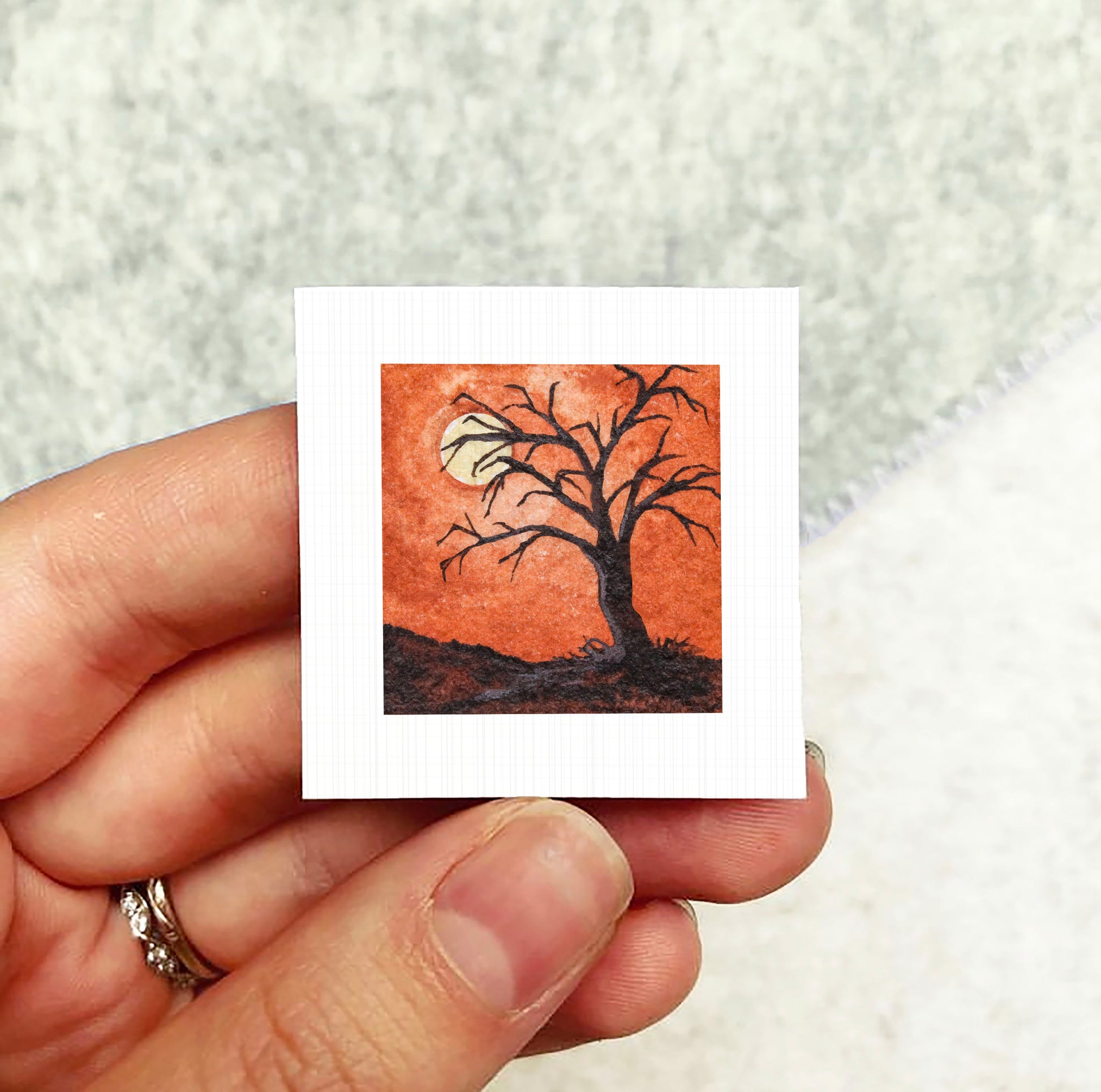 A hand holding a small square artwork depicting a tree with a full moon in the background.