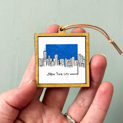 A hand holding a small wooden frame with a picture of a city skyline and the text "New York City" written on it.