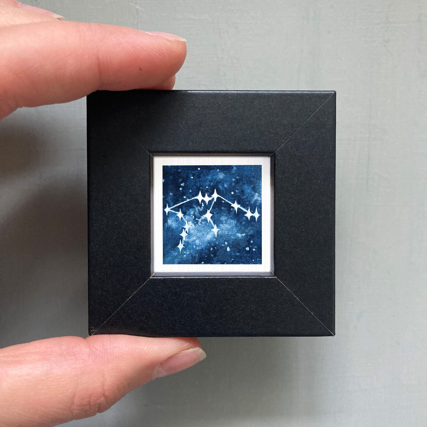 A hand is holding a small black square frame containing a framed image of a starry night sky.