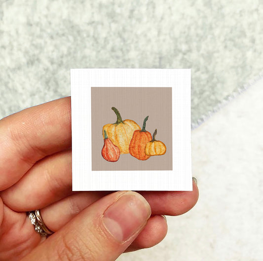 A hand holding a small square card with an illustration of three pumpkins on it.