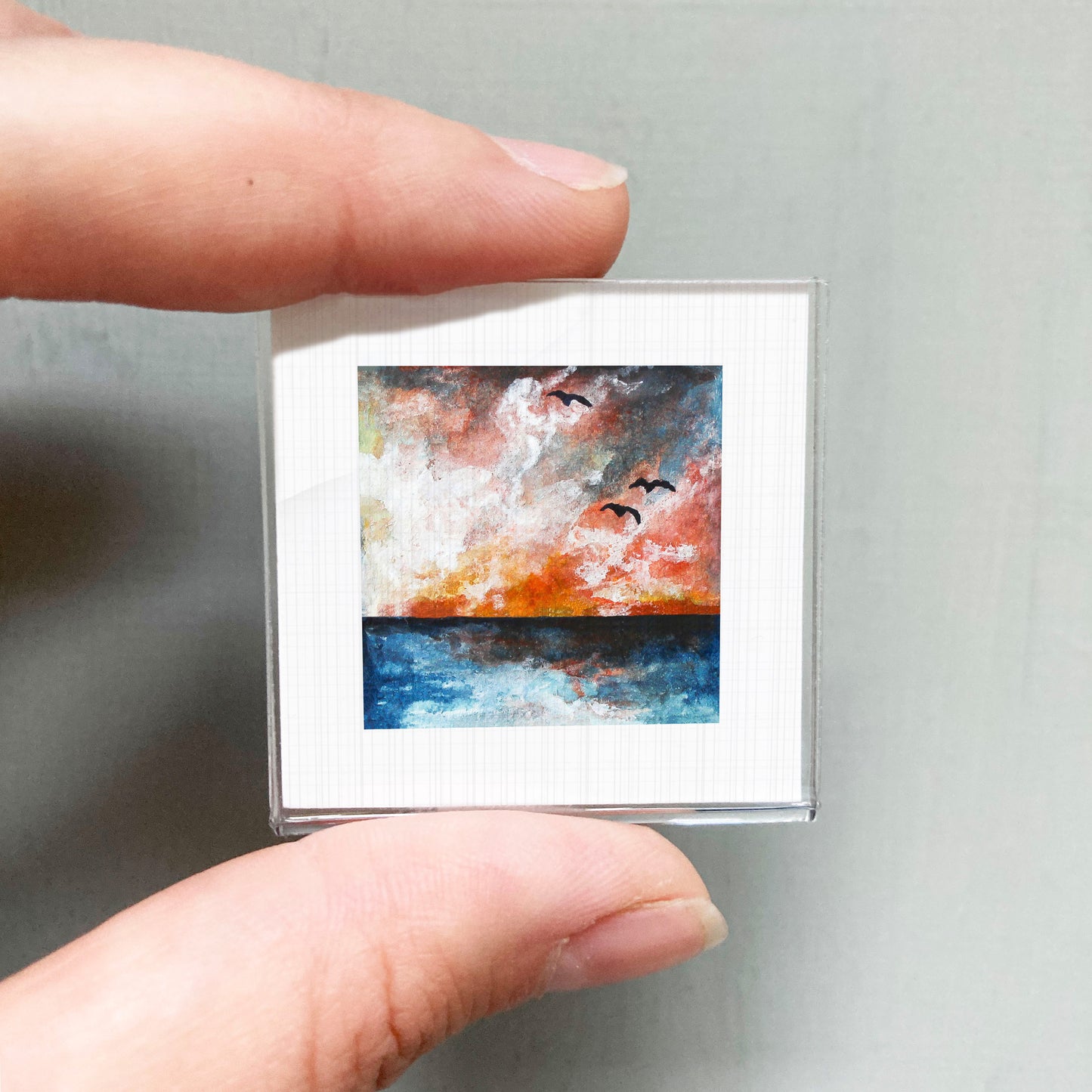 A person's hand is holding a small square picture of a painting depicting a sunset over a body of water.