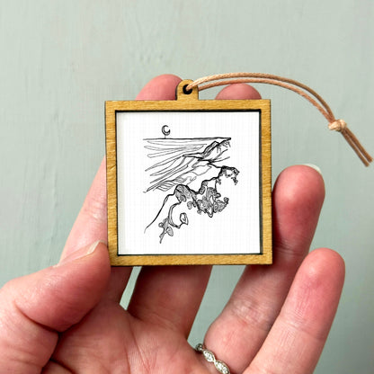 A hand holding a small wooden frame containing a black and white illustration of a bird or bird-like creature.