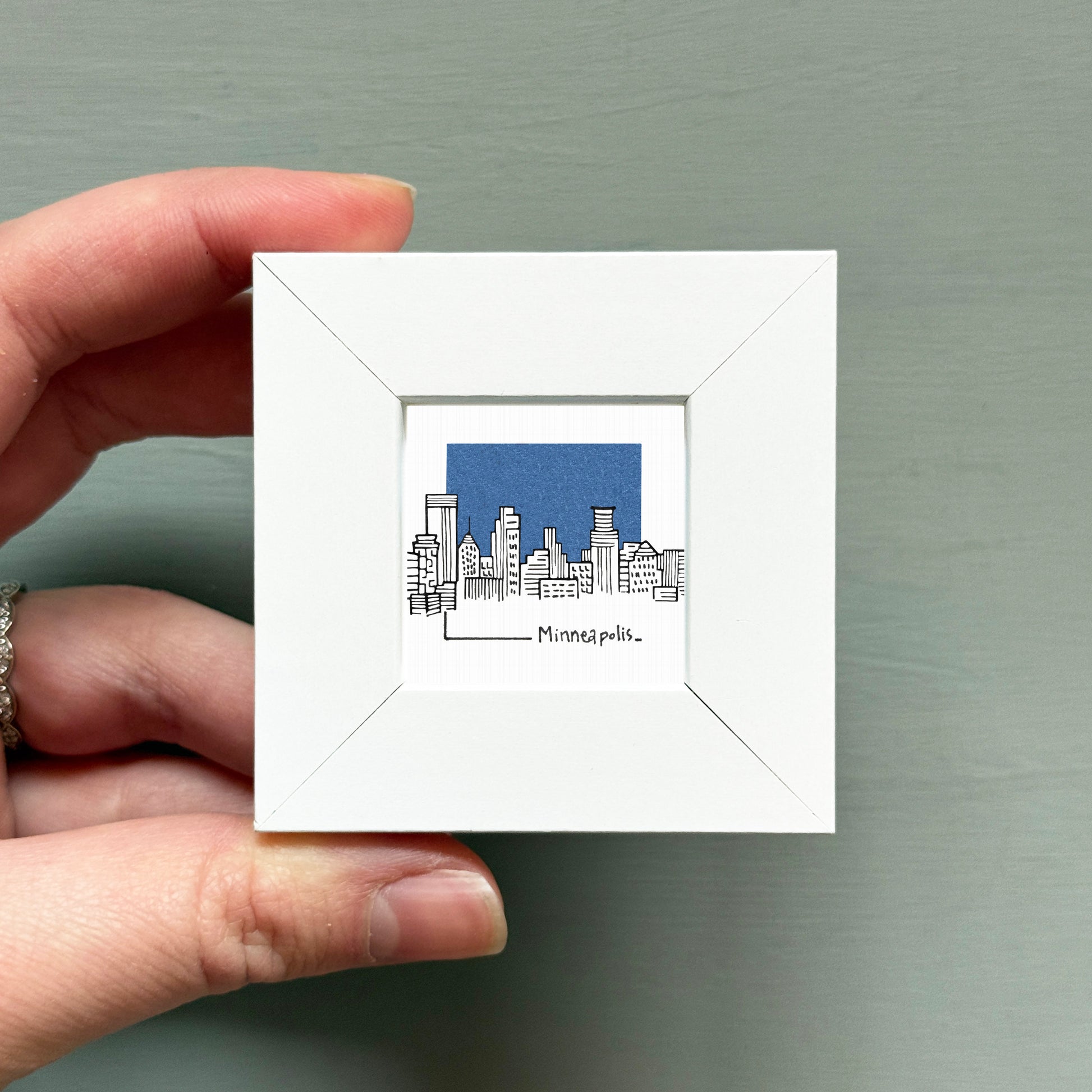 A hand is holding a small white picture frame containing an illustration of the Minneapolis skyline.