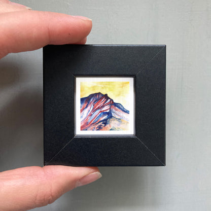 A person's hand is holding a small, square-shaped black frame containing a vibrant, colorful painting of a mountain landscape.
