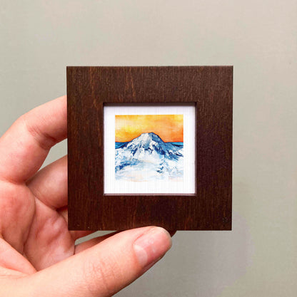 A hand is holding a small, square-shaped wooden frame containing a watercolor painting of a mountain landscape.