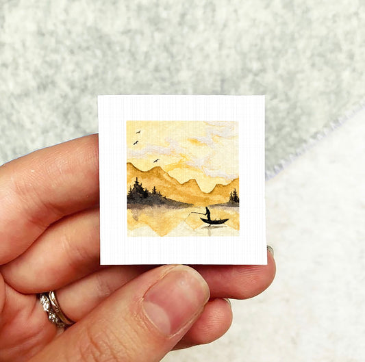 A hand holding a small square painting depicting a person in a boat on a lake, with mountains in the background.