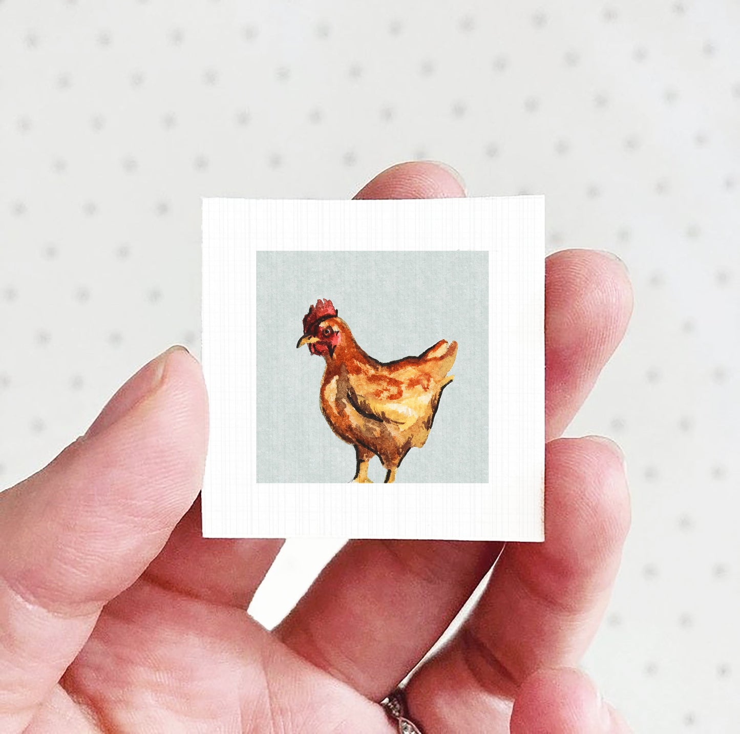 A hand holding a small square picture of a chicken against a white background.
