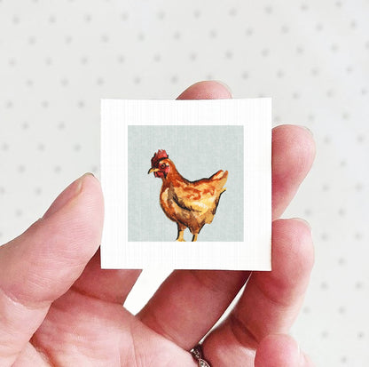 A hand holding a small square picture of a chicken against a white background.