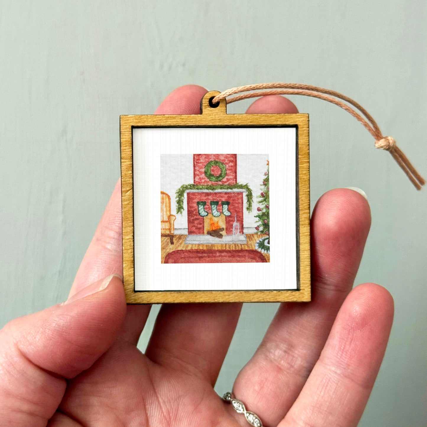A hand holding a small wooden picture frame containing a Christmas-themed image of a fireplace with stockings hanging above it.