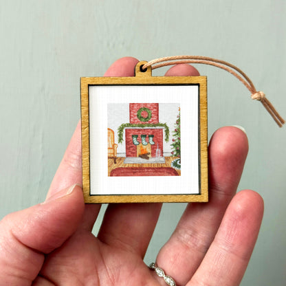 A hand holding a small wooden picture frame containing a Christmas-themed image of a fireplace with stockings hanging above it.